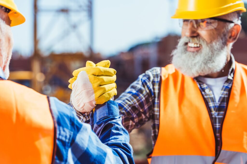 Smiling construction worker in protective uniform shaking hands at toolbox talk with colleague