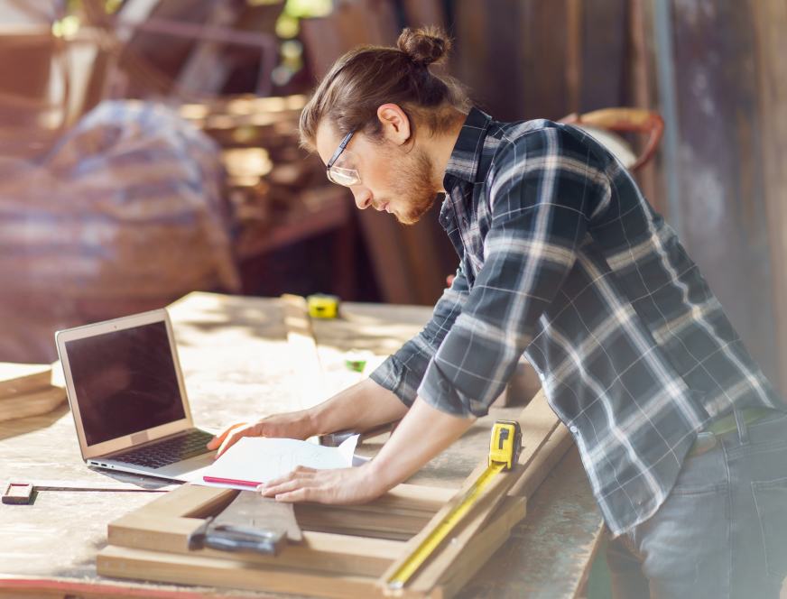 young hipster carpenter man working with computer laptop in workshop . craftsman wearing safety glasses checking order of clients or learning online