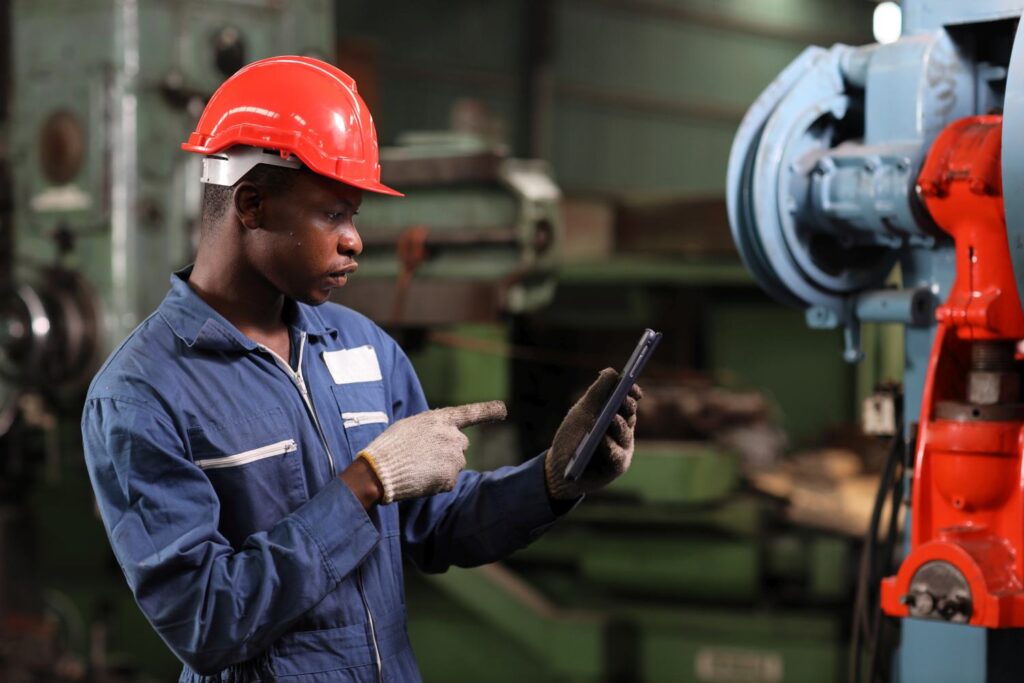Technician engineer and worker in protective uniform standing and using tablet while controlling operation or checking industry machine process with hardhat at heavy industry manufacturing factory