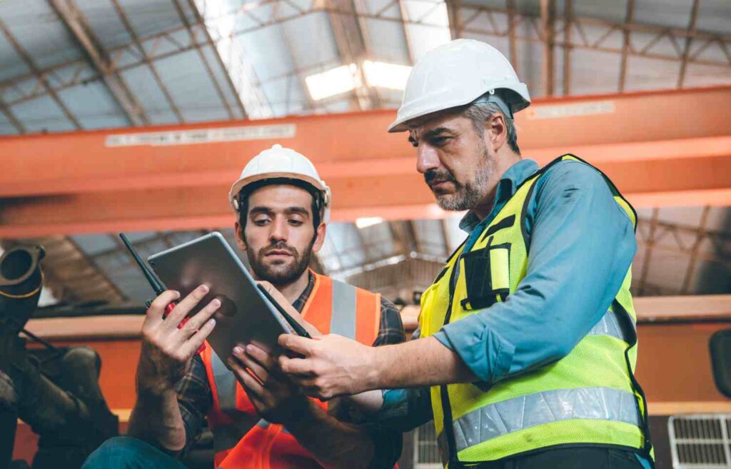 Two senior and young male Asian engineers in safety vest and jacket with hardhat and helmet working in warehouse and factory on a machine while fixing and inspecting equipment